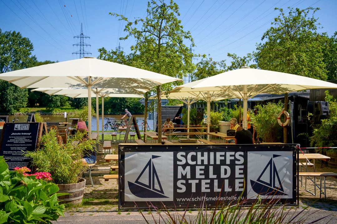 Der Eingang der Schiffsmeldestelle mit Banner, Sonnenschirmen und Bierzeltgarnituren am Mainufer