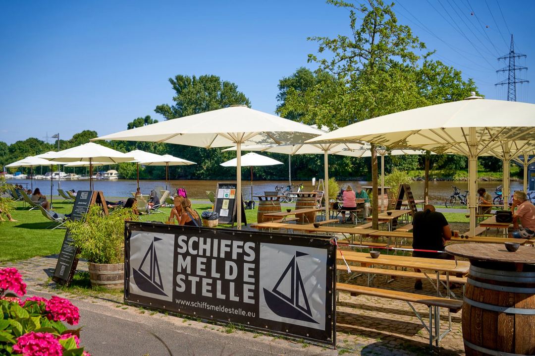 Bierzeltgarnituren und Sonnenschirme der Schiffsmeldestelle, dahinter Liegestuehle auf der Wiese und der Main